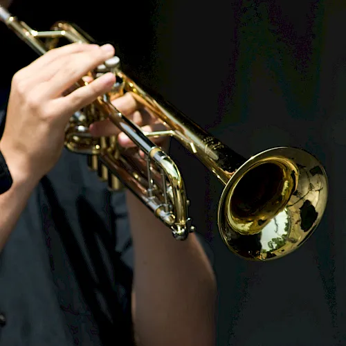 A person plays a shiny gold trumpet on stage, holding it up toward the camera with fingers pressing the valves.