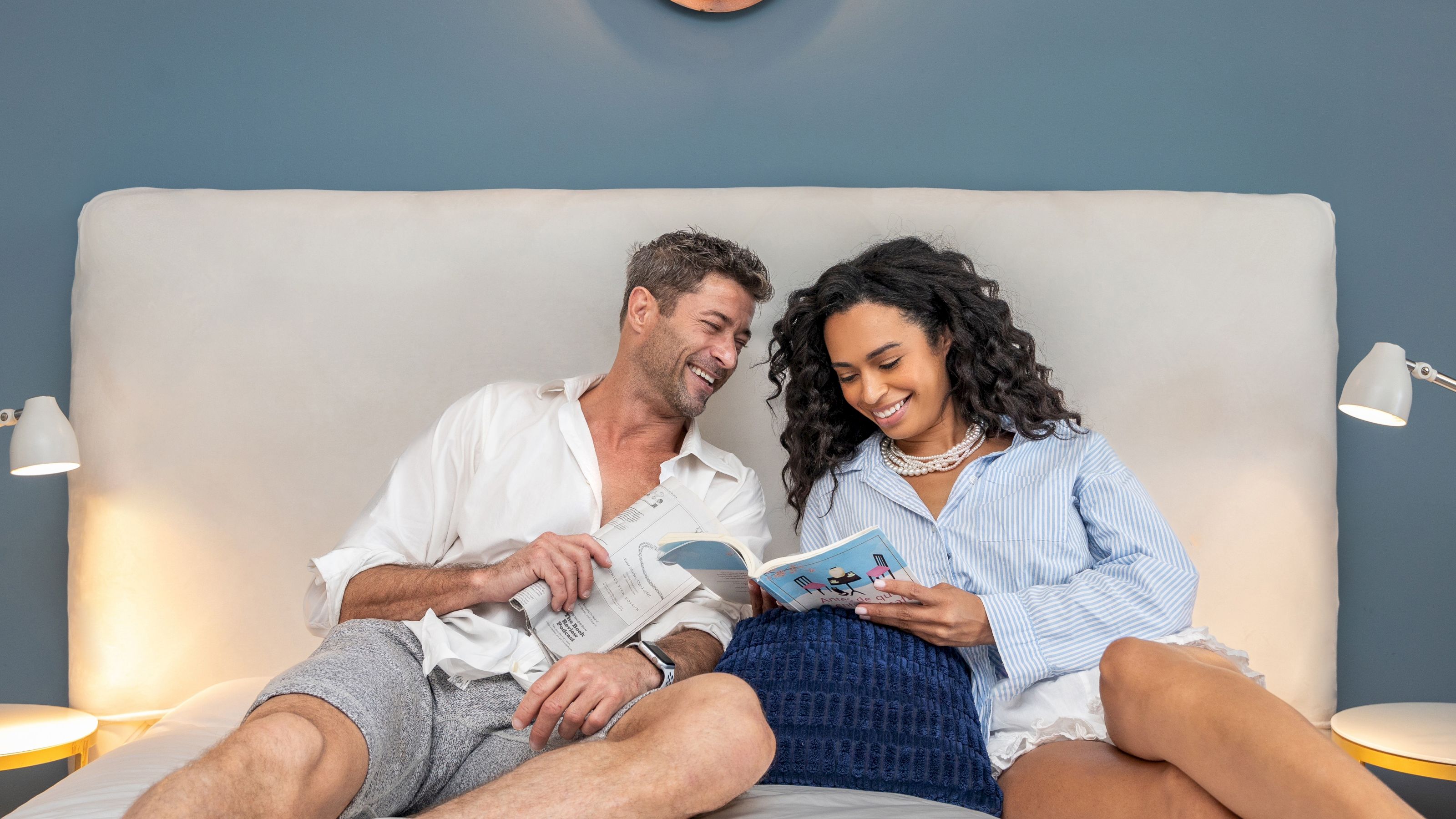 A couple reading in bed with a blue headboard and white lamps.