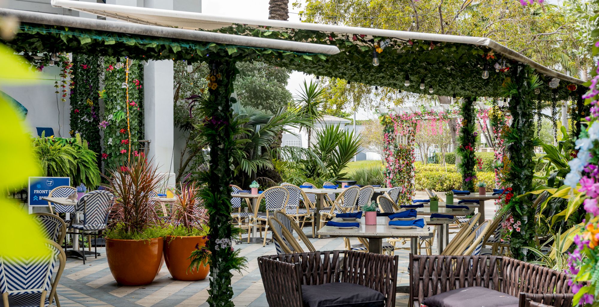 A shady outdoor cafe with vines-covered arches, wicker chairs, tables, potted plants, and a serene, plant-filled dining area.