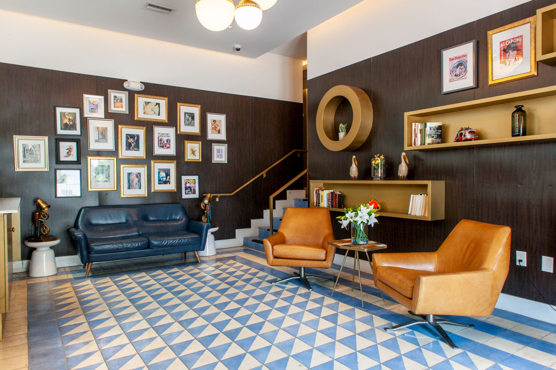 A stylish lounge with leather chairs, a blue sofa, geometric rug, wall art gallery, shelves, and pendant light.