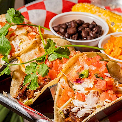 The image shows a tray with tacos topped with cilantro and tomatoes, served with black beans, rice, and corn on the cob.