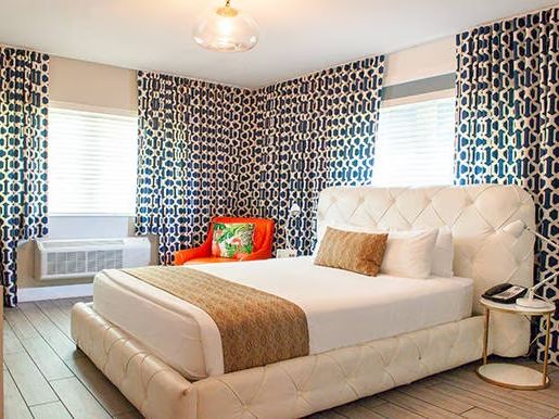 A stylish bedroom with a white bed, patterned blue curtains, an orange chair, and a side table, creating a modern, cozy ambiance.