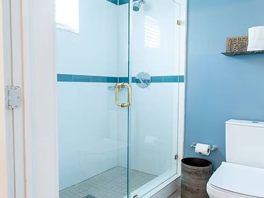 A bathroom with a glass shower, toilet, toilet paper holder, and towel shelf against a blue wall.