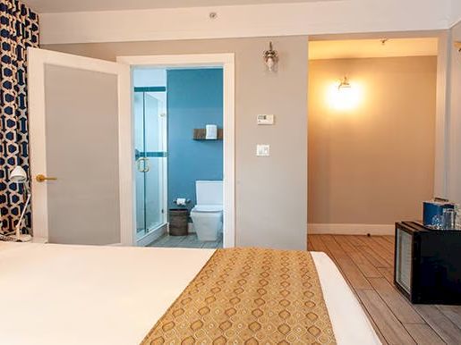 A hotel room with a bed, bathroom with a glass shower door, decorative wallpaper, and modern furnishings, including a mini fridge.