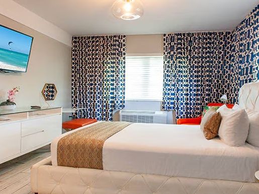 A modern bedroom with a TV, white furniture, patterned curtains, and a neatly made bed with white and gold bedding ends the look.