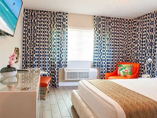 A hotel room with a bed, patterned curtains, wall-mounted TV, dresser, orange chair, and decorative pillows completes the scene.