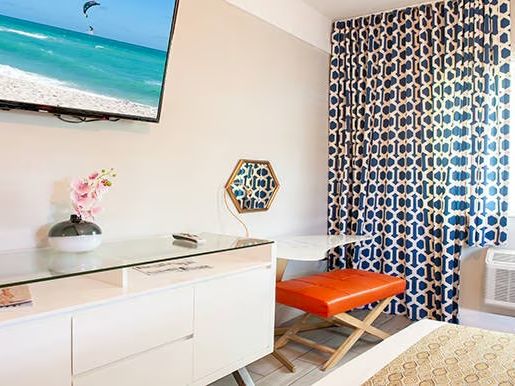 A modern room with a wall-mounted TV, white dresser, orange stool, blue patterned curtains, and decorative items.