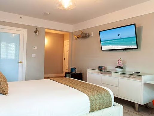 A modern hotel room features a bed, TV showing a beach scene, dresser, and a table with a lamp, in a neat and cozy setting.
