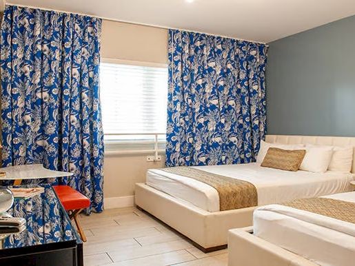 The image shows a hotel room with two beds, a TV, a table, blue floral curtains, and modern decor.
