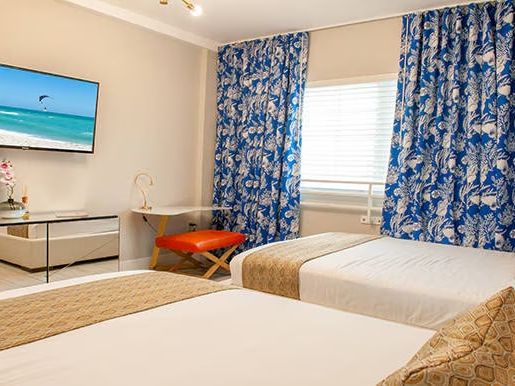 A hotel room with two beds, blue patterned curtains, a wall-mounted TV, and a small desk with a chair and decorative items.