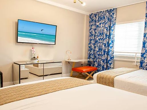 The image shows a hotel room with two beds, a TV, a dresser, and blue-patterned curtains, creating a bright and cozy atmosphere.