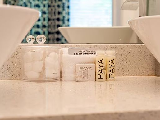 The image shows a bathroom counter with two white sinks, cotton balls in a jar, and Paya-branded toiletries, including soap and lotion.