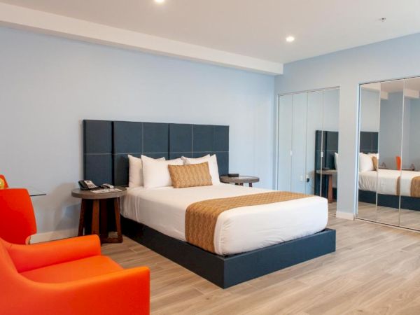 A modern hotel room with a bed, orange chairs, large mirror, and minimalistic decor, featuring wooden flooring and blue walls.