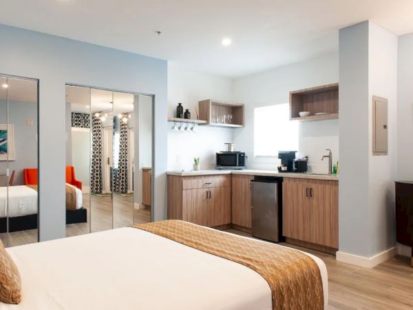The image shows a modern hotel room with a bed, kitchenette, and mirrors, featuring wood accents and neutral tones in the decor.