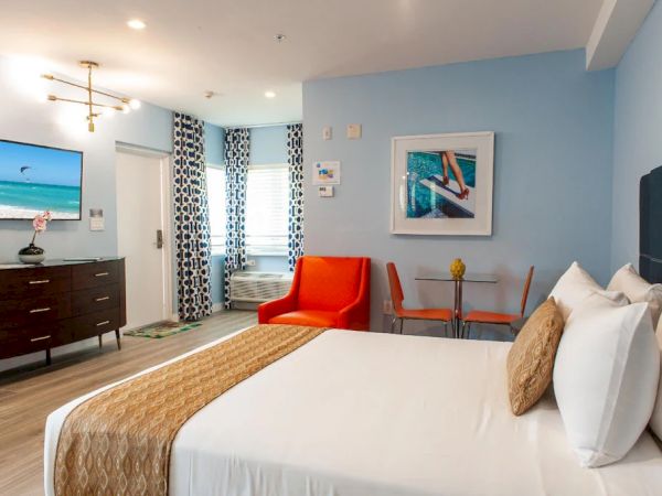 A hotel room with a bed, orange chair, TV, dresser, table, and beach artwork. Walls are light blue, and curtains have a geometric pattern.