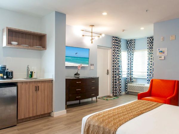 The image shows a hotel room with modern decor, featuring a bed, orange chair, TV, kitchenette, and a window with patterned curtains.