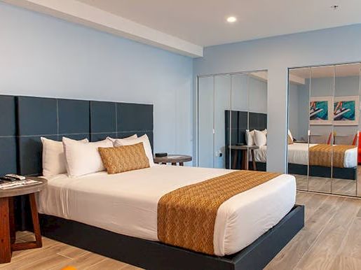 A modern hotel room features a bed, pillows, side tables, mirrored closet, wall art, and a red chair on wooden flooring.