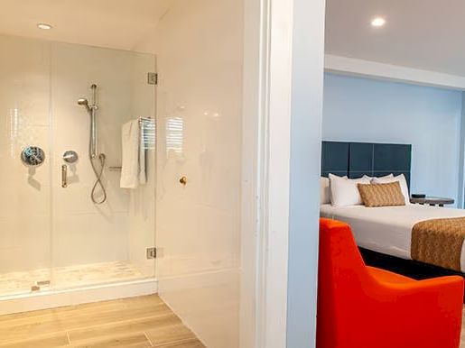 The image shows a modern hotel room with a bed, orange chair, and adjacent glass shower enclosure in the bathroom.