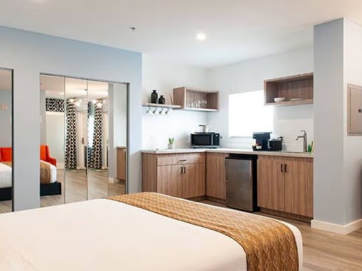 A modern hotel room with a bed, kitchenette, mirrored closet, TV, and wood flooring.
