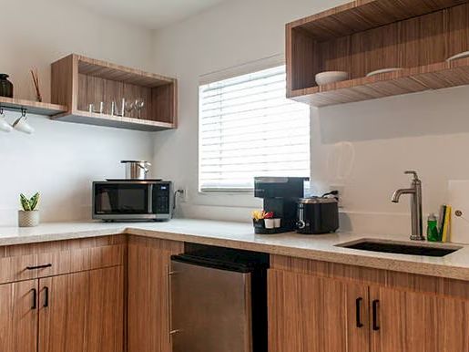 A modern kitchen with wooden cabinets, open shelves, a microwave, a coffee maker, a small fridge, a sink, and a window with blinds.