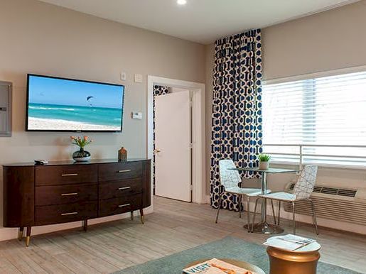 A modern living area with a TV, wooden dresser, round table with chairs, patterned curtains, and coastal decor accents the relaxed ambiance.