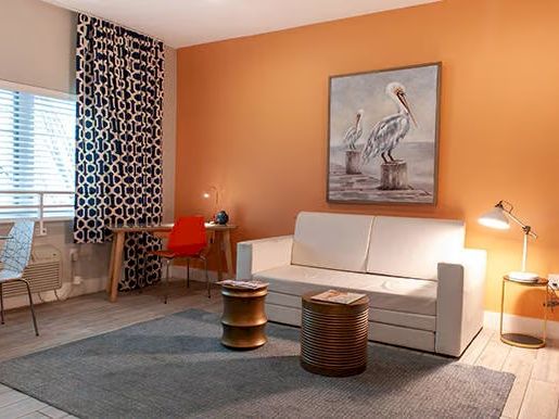A modern living room with orange walls, white sofa, pelican artwork, wooden stools, patterned curtains, and a small dining area on the left.