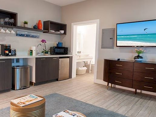 A modern room with a kitchenette, wall-mounted TV, and a dresser. It has a beach scene on the TV and a cozy, inviting atmosphere.