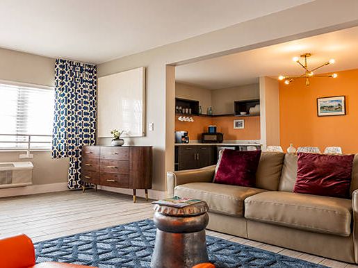 A modern living room with a beige sofa, orange armchairs, blue rug, and an open kitchen area with an orange accent wall.