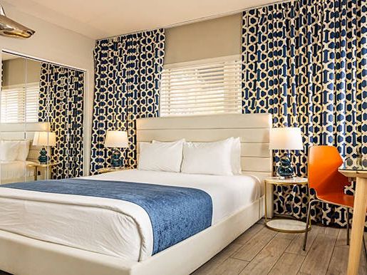 A modern bedroom with patterned curtains, a white bed with a blue throw, two lamps, a mirrored wall, and an orange chair at a desk.