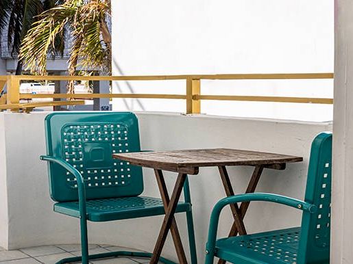 Two teal chairs and a wooden table on a balcony with yellow railing and a palm tree in the background.