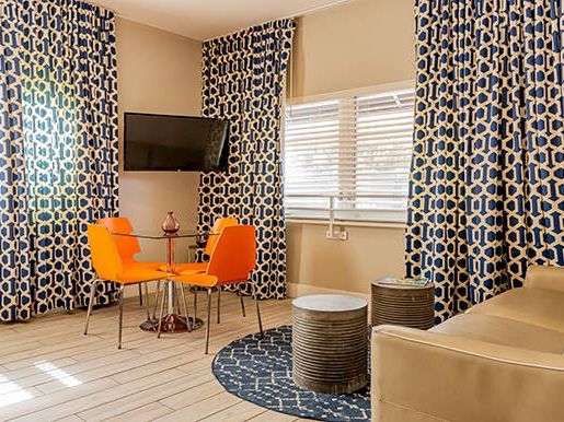 A modern room with patterned curtains, a flat-screen TV, an orange dining set, two small tables, and a beige sofa on a wooden floor.