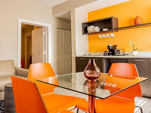 A modern kitchen with orange accents, glass table, and sink area, featuring orange chairs and shelves with dishes.