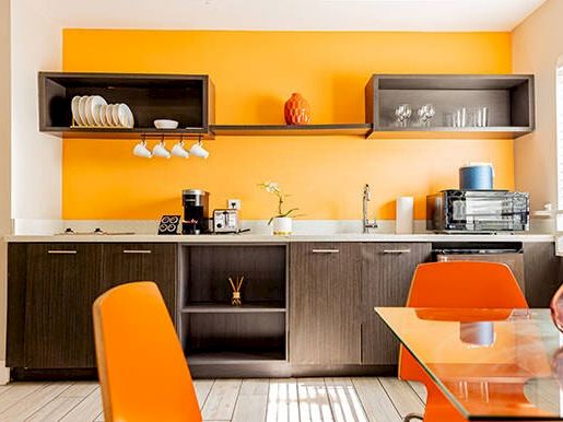 A modern kitchen with orange chairs, a glass table, dark cabinets, and a bright orange wall with shelves and various kitchen items arranged.