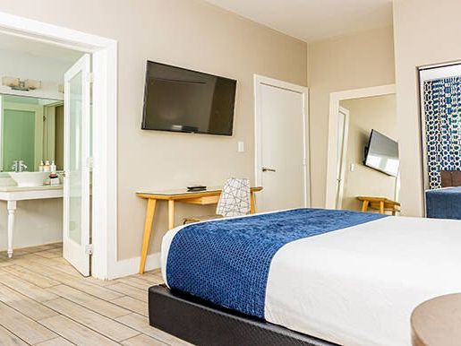 The image shows a modern hotel room with a bed, TV, desk, bathroom, and a mirror, featuring blue and white decor.