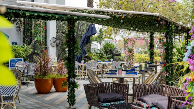 A pergola with hanging greenery and flowers covers an outdoor dining area with tables and chairs.