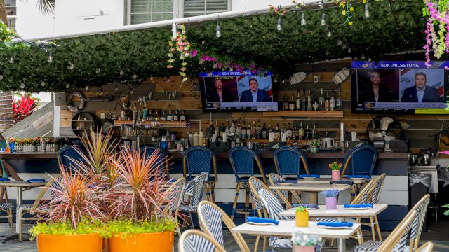 A bar with tables, chairs, TV screens, and a counter with bottles and plants.