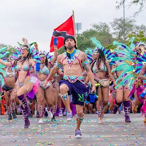 A group of dancers in colorful, feathered costumes perform energetically in a parade, with a red flag and palm trees in the background.