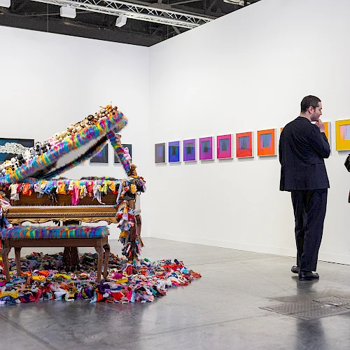 An art gallery features a colorfully decorated piano and visitors observing framed abstract artworks on the walls.