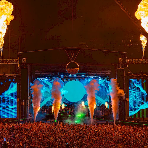 A large crowd at an outdoor concert with vibrant blue lighting, smoke effects, and fire bursts on stage create an electrifying atmosphere.