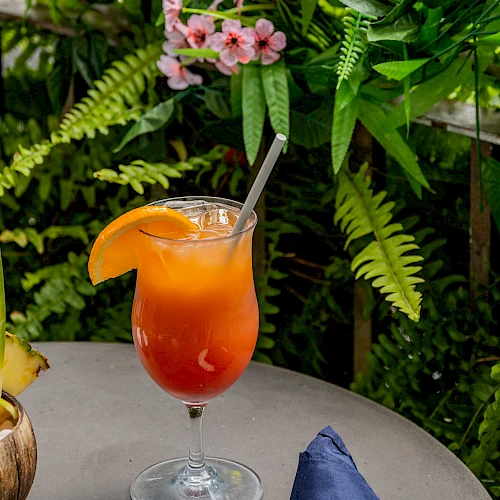 A tropical cocktail with an orange slice garnish on a table, surrounded by lush green ferns and pink flowers, next to a blue napkin.