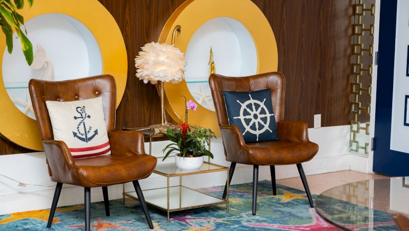 Two leather armchairs with nautical-themed pillows sit in a lobby area with a coffee table and lamp.