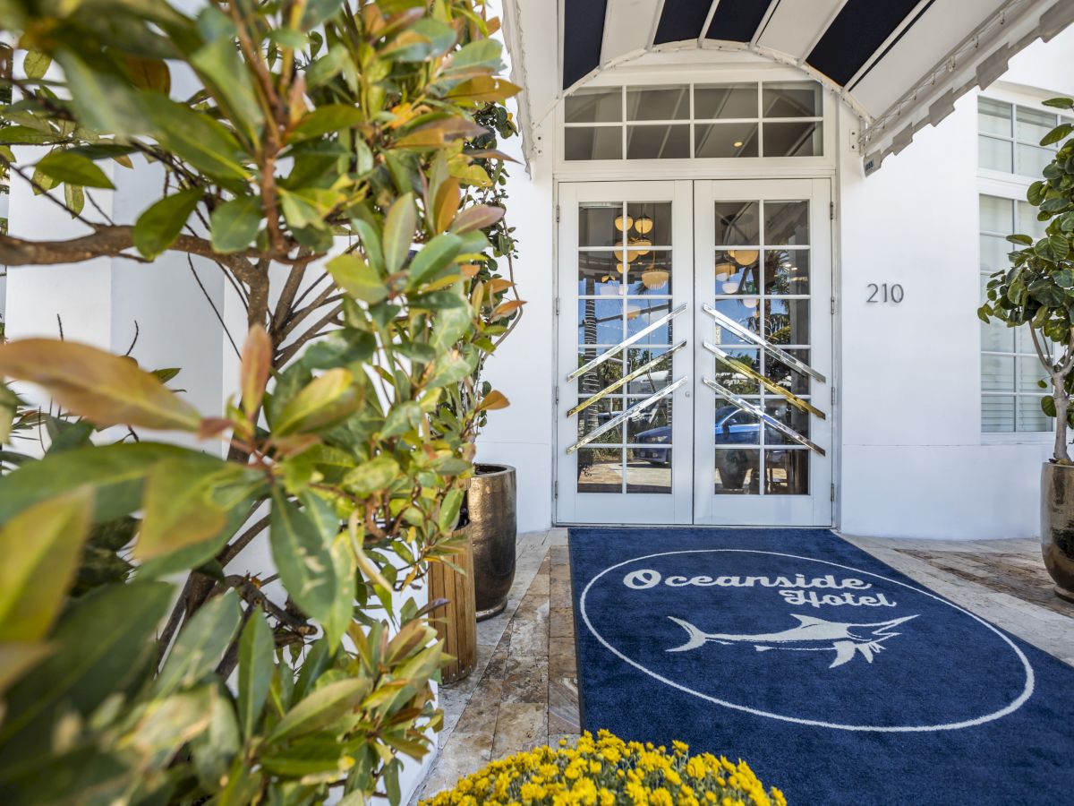 Entrance of the Oceanside Hotel with potted plants, a blue mat featuring a fish design, and glass double doors at address 210.