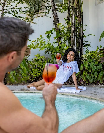 Two people by a pool, one man raising a drink toward the camera while a woman sits on the pool deck smiling with a colorful cocktail.