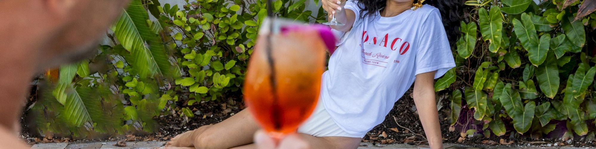 Two friends by a pool: a man in foreground raises a orange cocktail while a woman lounges on the edge, smiling behind tropical plants.