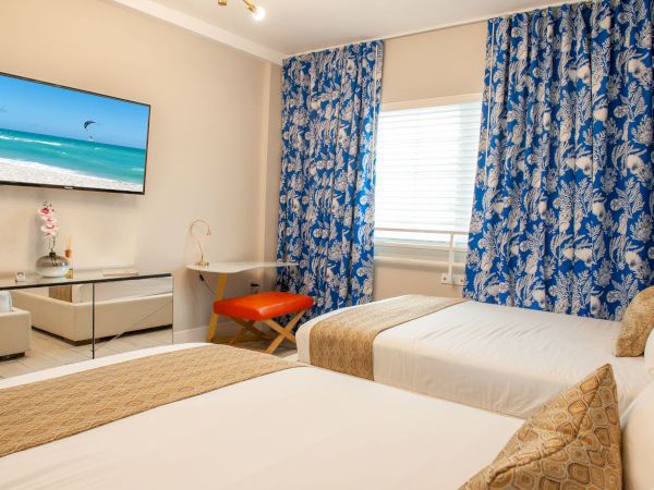 A bright hotel room with two single beds, a wall-mounted TV, blue patterned curtains, a small desk, and an orange chair by the window.