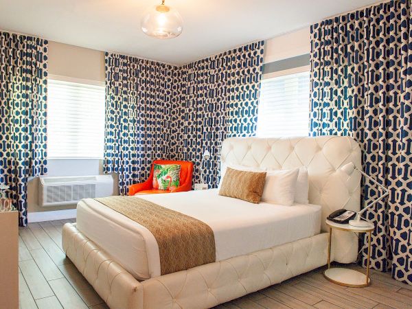 A modern hotel bedroom with a tufted white bed, bold geometric blue-pattern curtains, a small side table, and a window AC unit, bright and stylish.