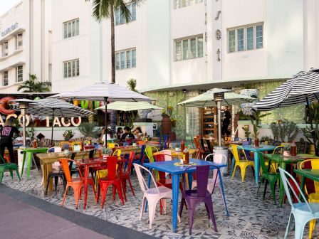 A sunny outdoor cafe with colorful plastic chairs and tables under umbrellas, in front of a white building with palm trees, bustling with people.
