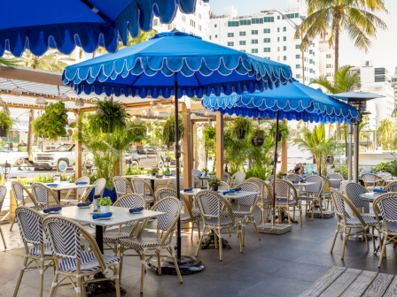 Outdoor cafe with blue striped umbrellas, wicker chairs, and tables under shade; palm trees and buildings in the background.