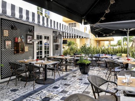 An outdoor cafe with black-and-white patterned walls, striped awnings, and numerous small tables and chairs on a tiled patio. The space looks cozy and shaded.