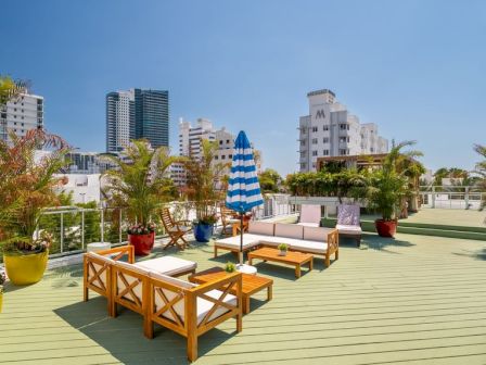 Rooftop terrace with wooden seating, potted plants, a blue striped umbrella, and city buildings in the background, sunny and calm.